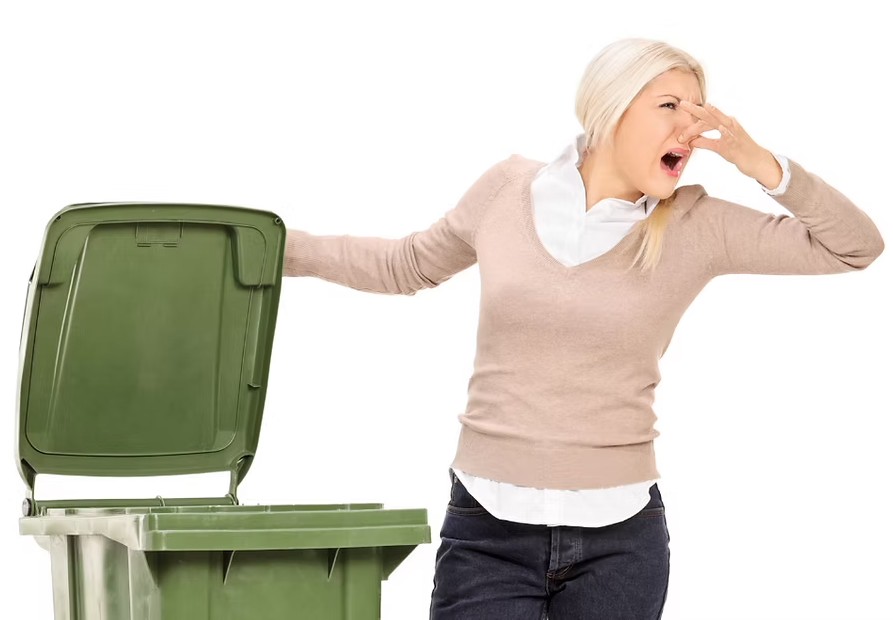 Woman holding her nose next to a smelly green trash bin